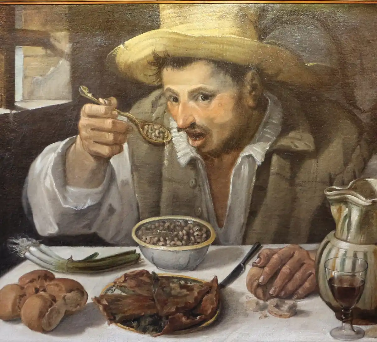 A man in a straw hat caught mid-spoonful over a bowl of beans, with spring onions, bread rolls and a jug on the white cloth before him.