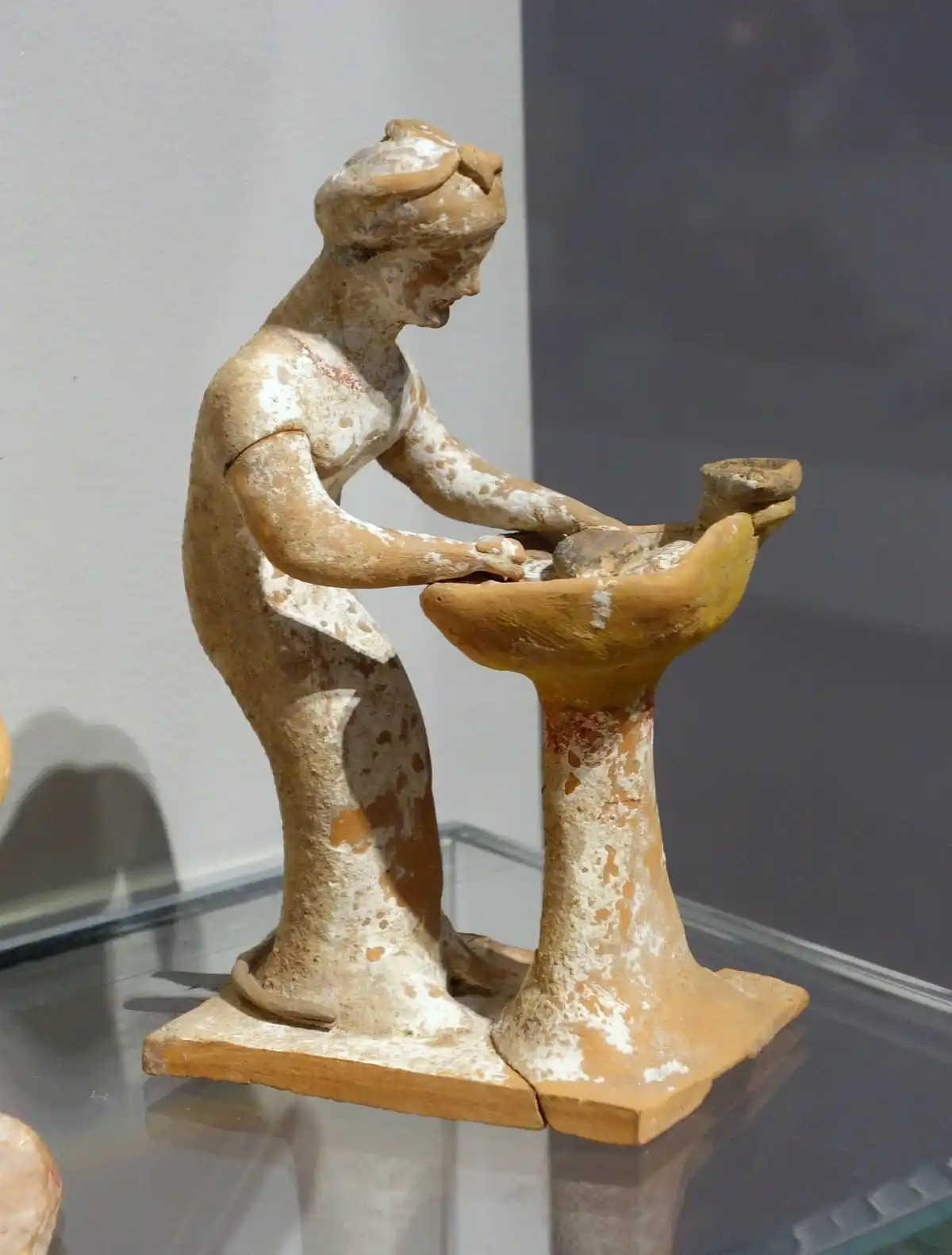 Greek terracotta figurine of a woman bent over a pedestal basin, kneading bread dough.