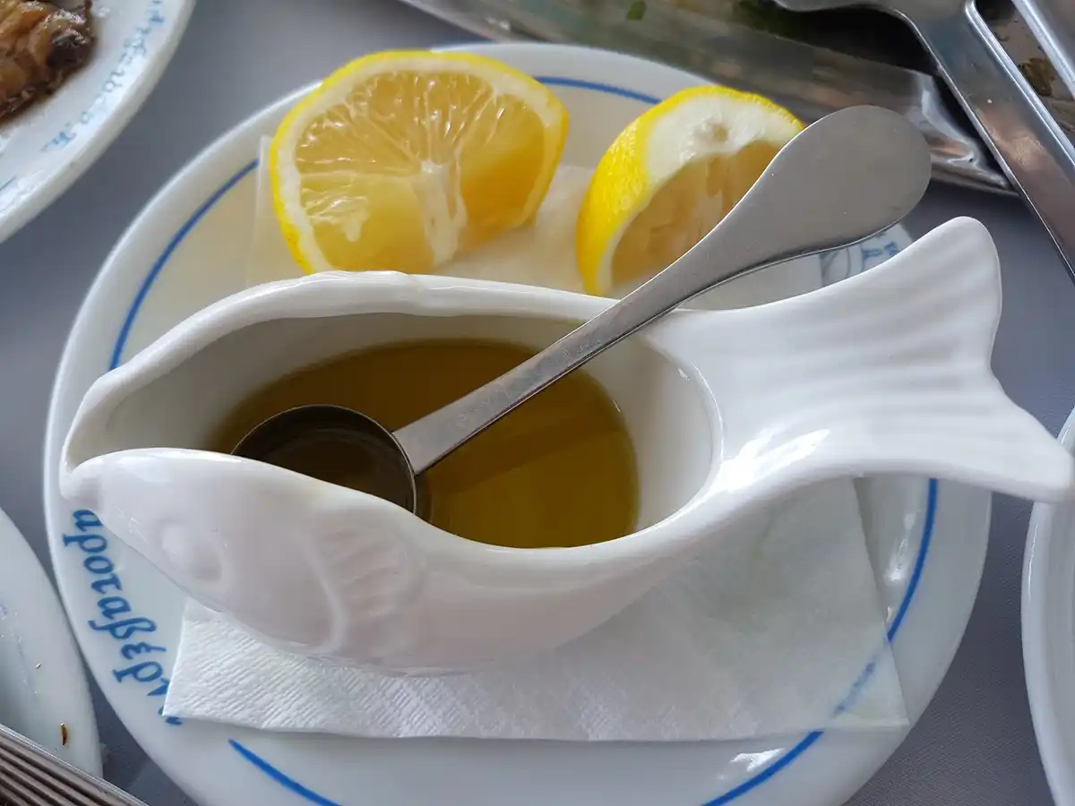 Golden ladolemono in a white fish-shaped sauce boat with a spoon, cut lemons beside it on a taverna table.