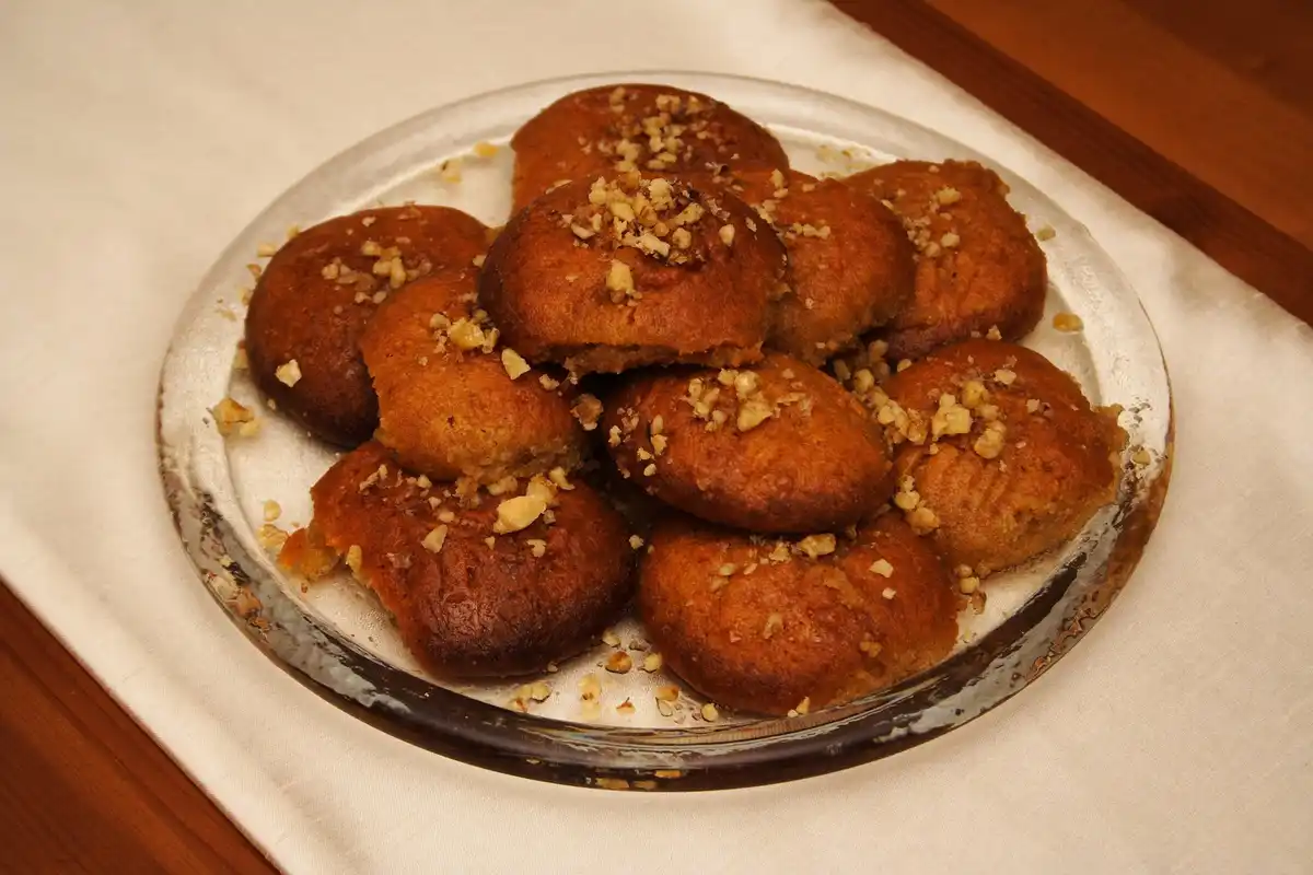 A pile of dark syrup-glazed melomakarona strewn with crushed walnuts on a glass plate over cream linen.