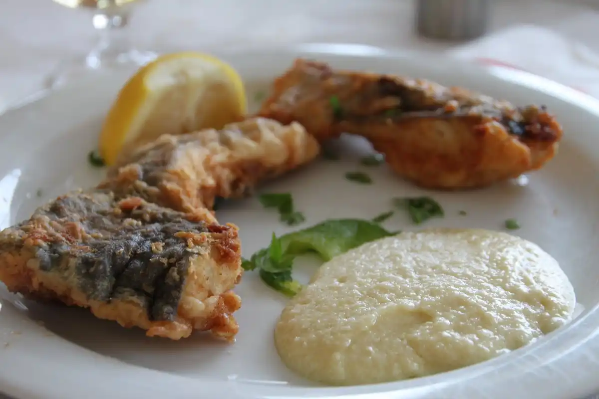 Golden battered fried cod with a lemon wedge and a dollop of pale skordalia on a white taverna plate, a wine glass behind.